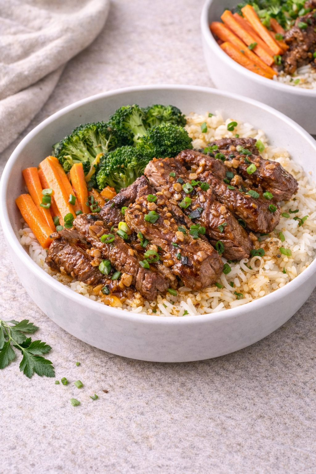 Bowl of white rice topped with sliced glazed beef, chopped scallions, and a sprinkle of sesame, plus broccoli and carrot sticks on the side.