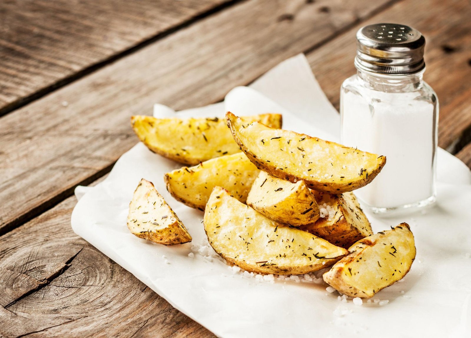 Crispy potato wedges sprinkled with herbs on a white parchment, with a salt shaker on a wooden table edge.