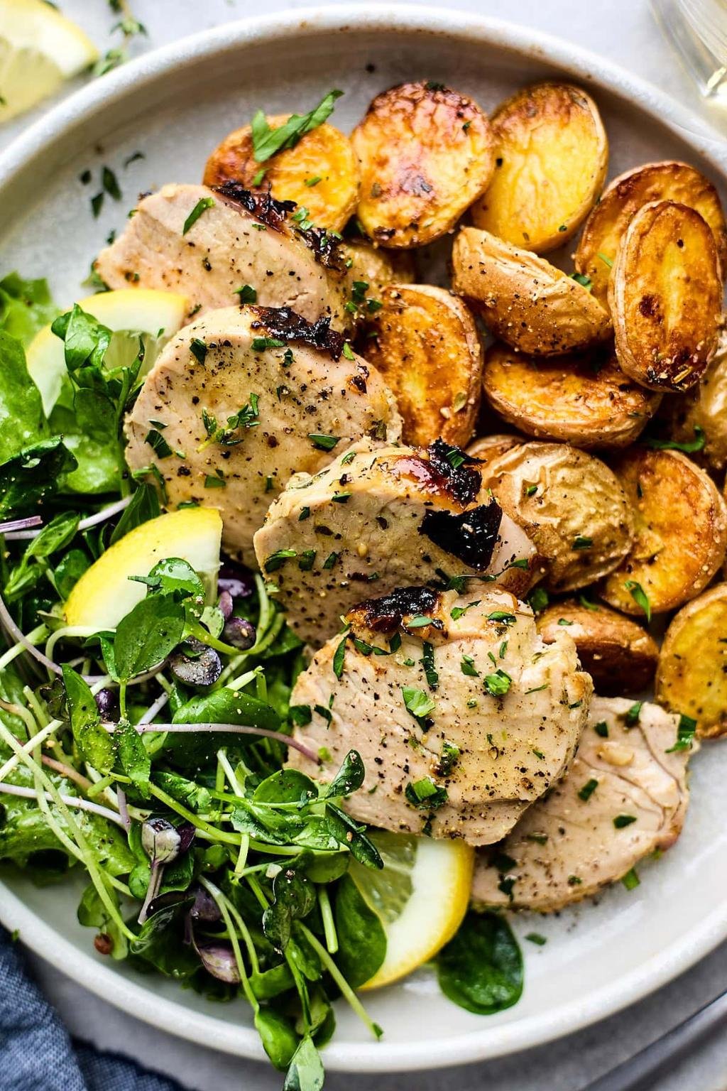 Sliced herb-seasoned chicken with roasted potatoes and a greens salad with lemon on a white plate.