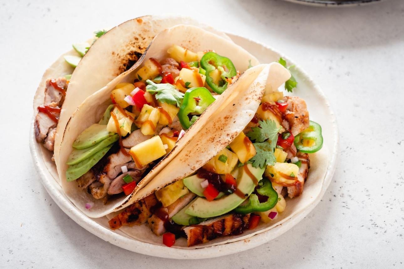 Two soft tacos filled with grilled pork, pineapple salsa, avocado, cilantro, onions, and jalapeño on a white plate.