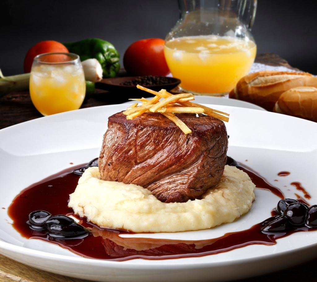 Beef tenderloin sitting on mashed potatoes with a dark red wine sauce around it and a crispy potato garnish on top, on a white plate.