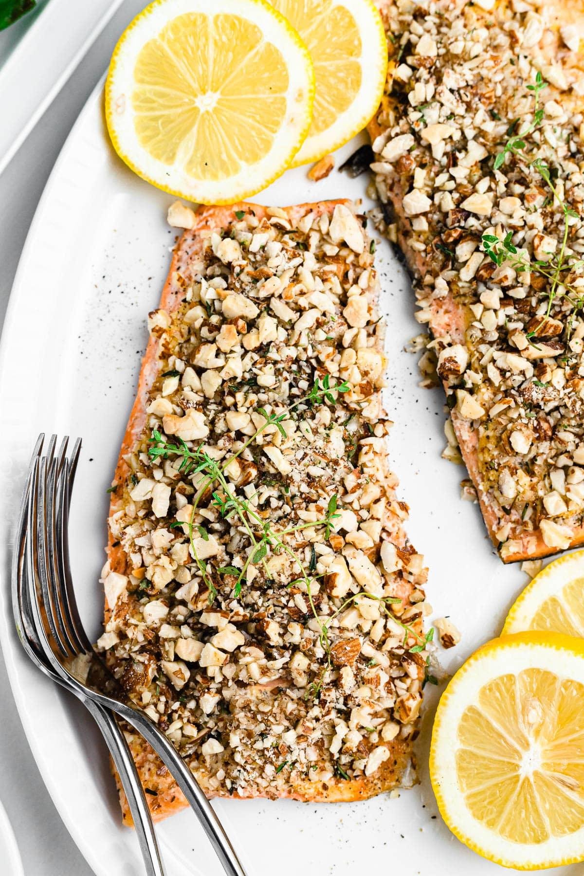 Salmon fillets crusted with chopped nuts and herbs on a white plate with lemon wedges nearby.