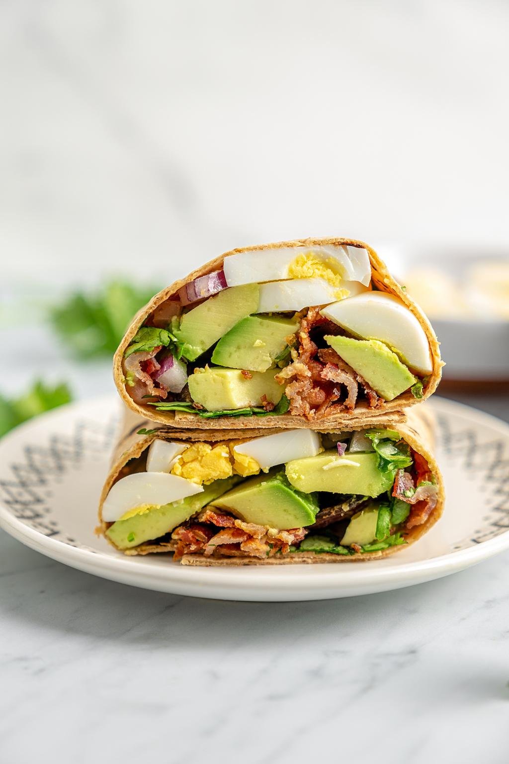 Two halves of a breakfast tortilla wrap filled with avocado slices, hard-boiled egg, red onion, greens and crispy bacon, on a plate.