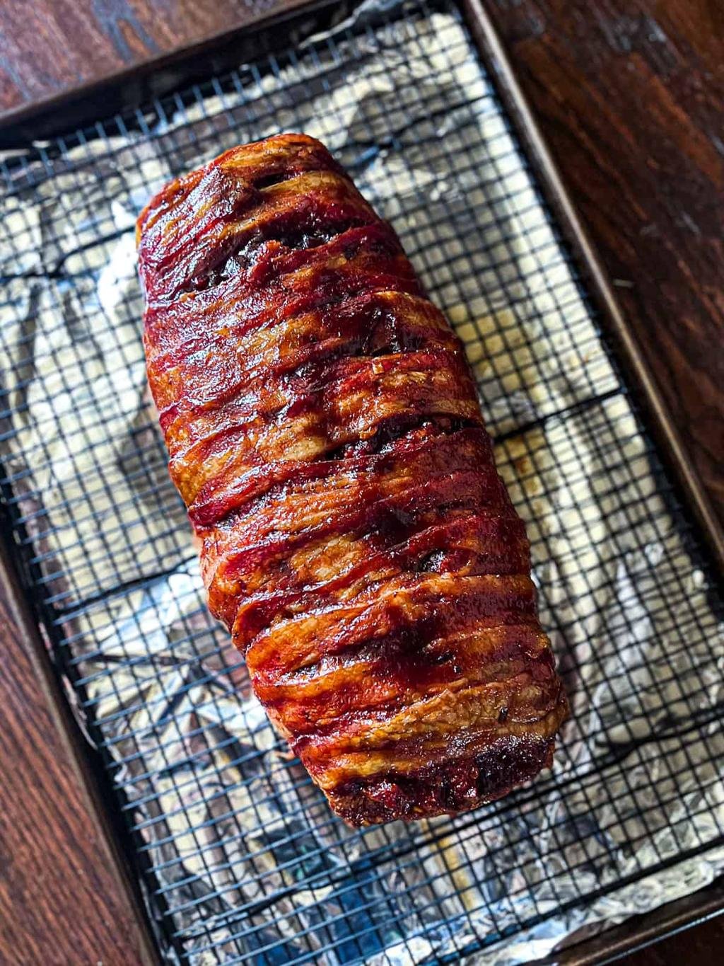Bacon-wrapped meat loaf or roast resting on a wire rack, caramelized and crispy edges visible.