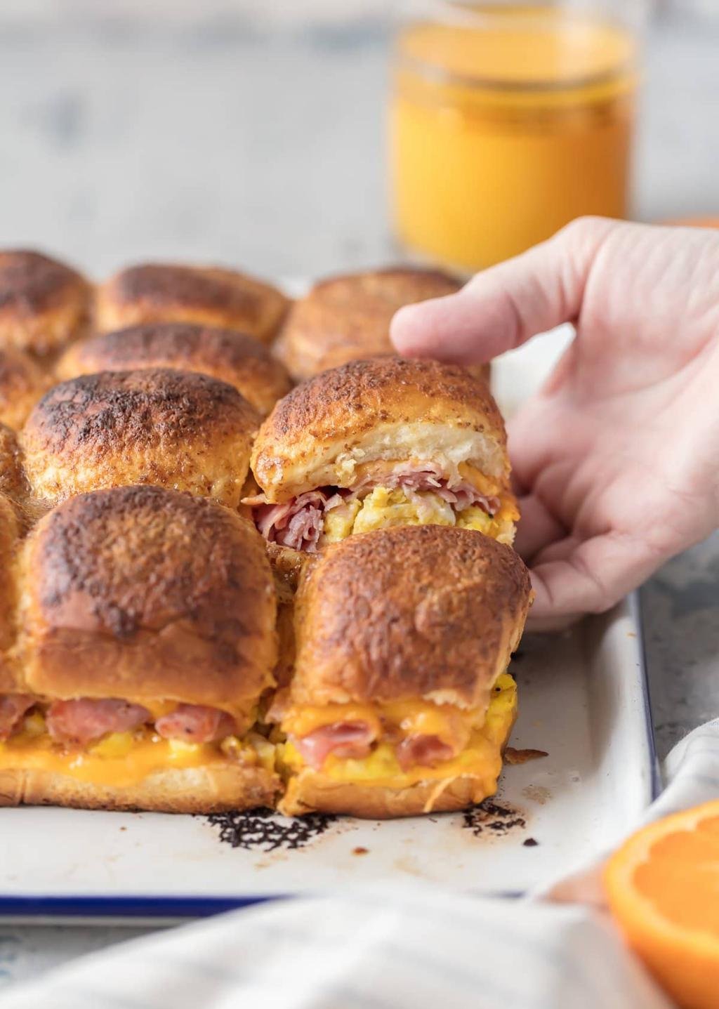 Close-up of ham and cheese sliders on a baking tray; a hand lifts one to show melted cheese inside, with a glass of orange juice in the blurred background.