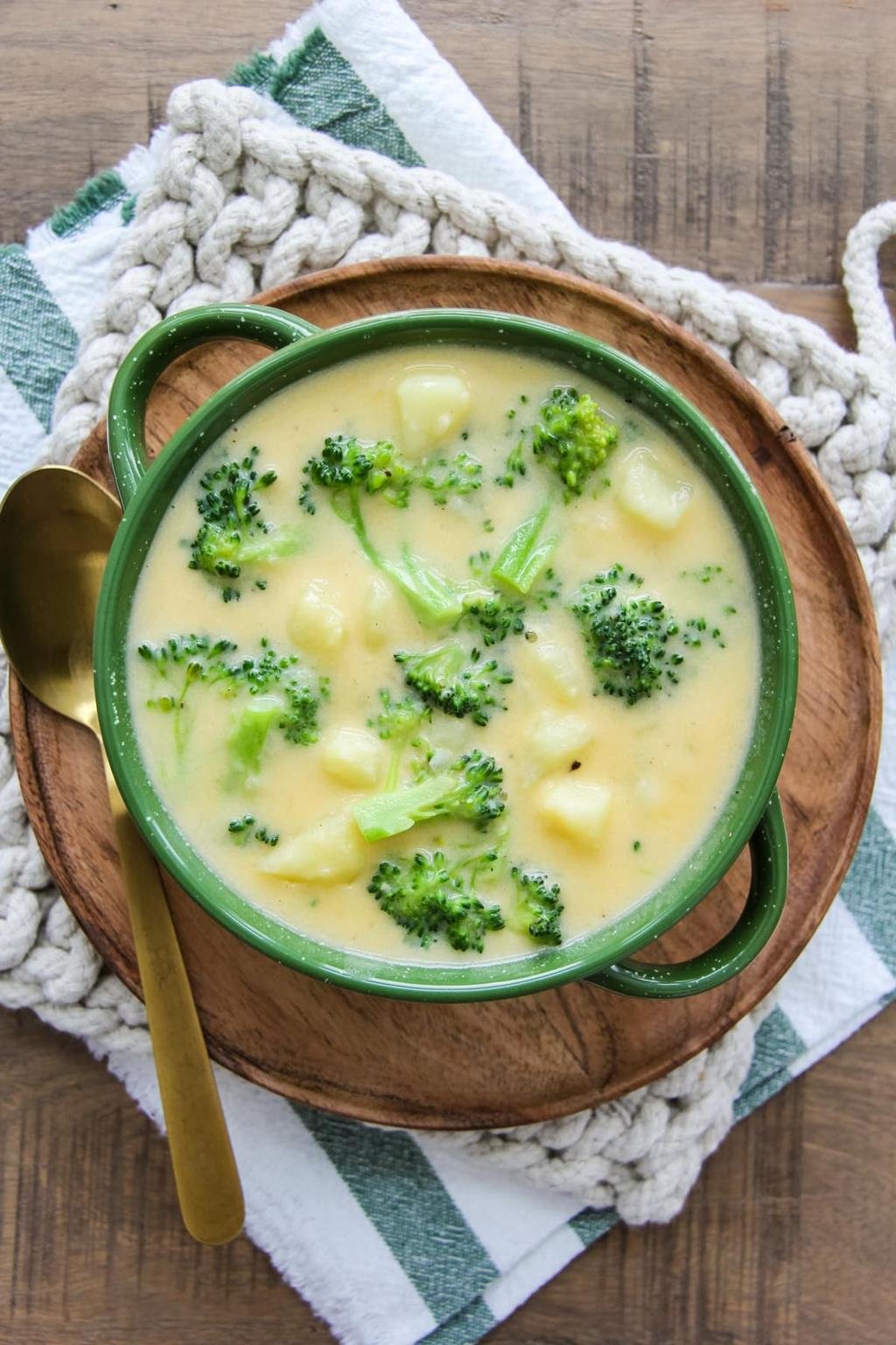 Green pot of creamy broccoli cheddar soup with potatoes, garnished with broccoli florets, on a wooden board with a gold spoon nearby.