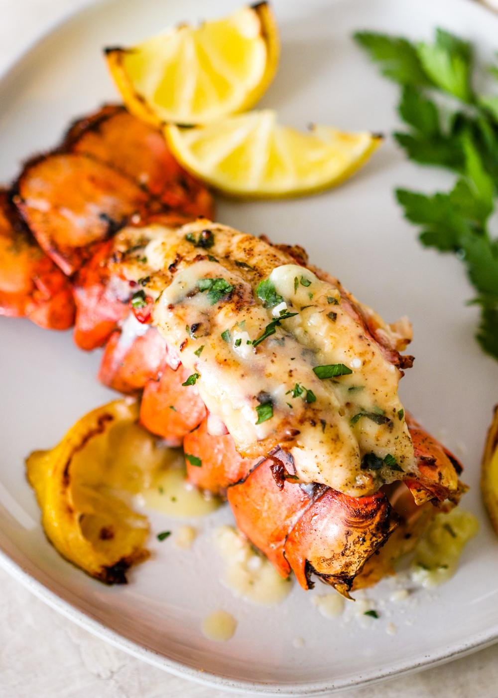 Lobster tail topped with melting herb-butter sauce and chopped parsley on a white plate, with lemon wedges nearby.