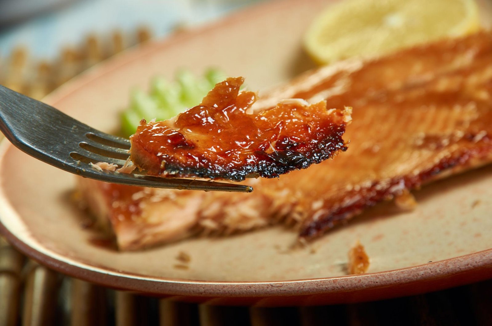 Close-up of a glazed barbecued rib on a fork, with a rack of ribs and a lemon wedge in the background on a plate
