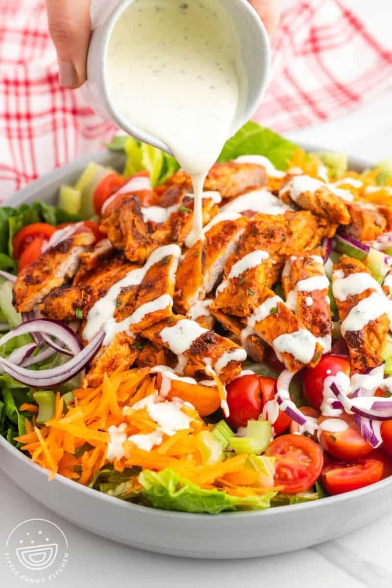 Salad bowl with lettuce, cherry tomatoes, shredded carrot, red onion, and sliced grilled chicken, as creamy dressing is poured on top.