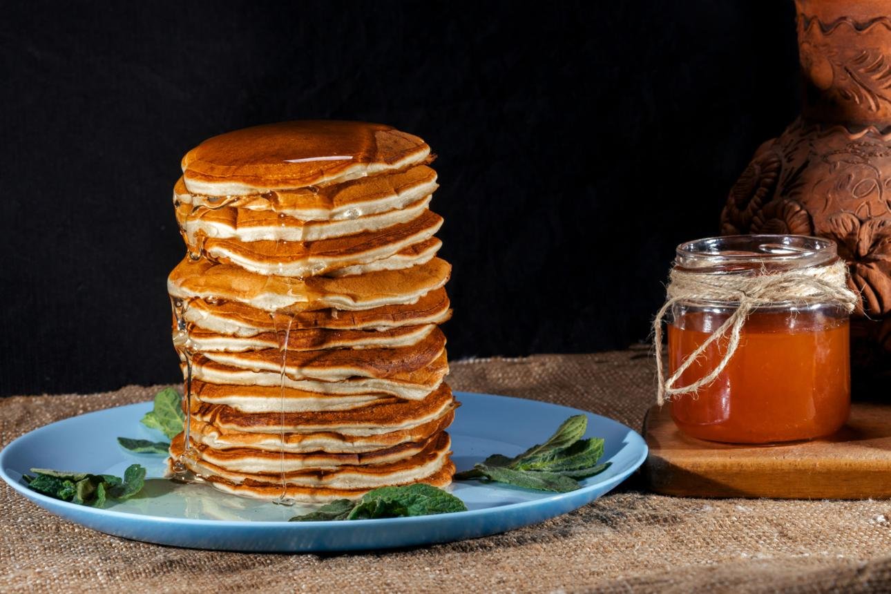Towering stack of pancakes with syrup on a blue plate, mint leaves nearby, and a honey jar tied with twine to the right.