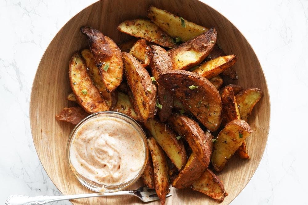 Crispy potato wedges on a wooden plate with a small bowl of creamy dipping sauce and a spoon nearby