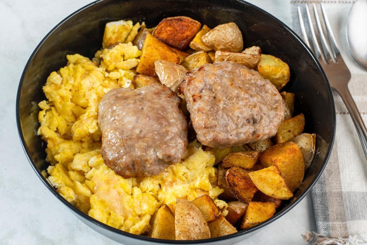 Black bowl with scrambled eggs, roasted potatoes, and two beef patties on top.