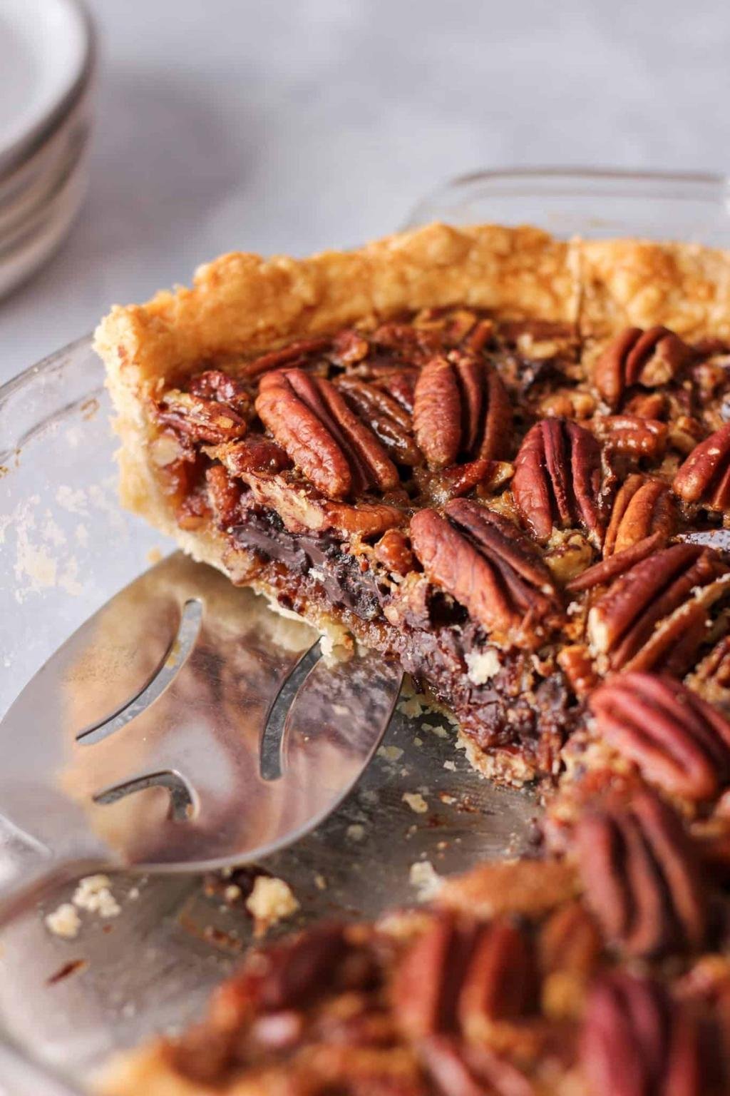 Close-up of a pecan pie with glossy pecan halves and a flaky crust, a slice removed and resting in a glass baking dish with a pie server nearby