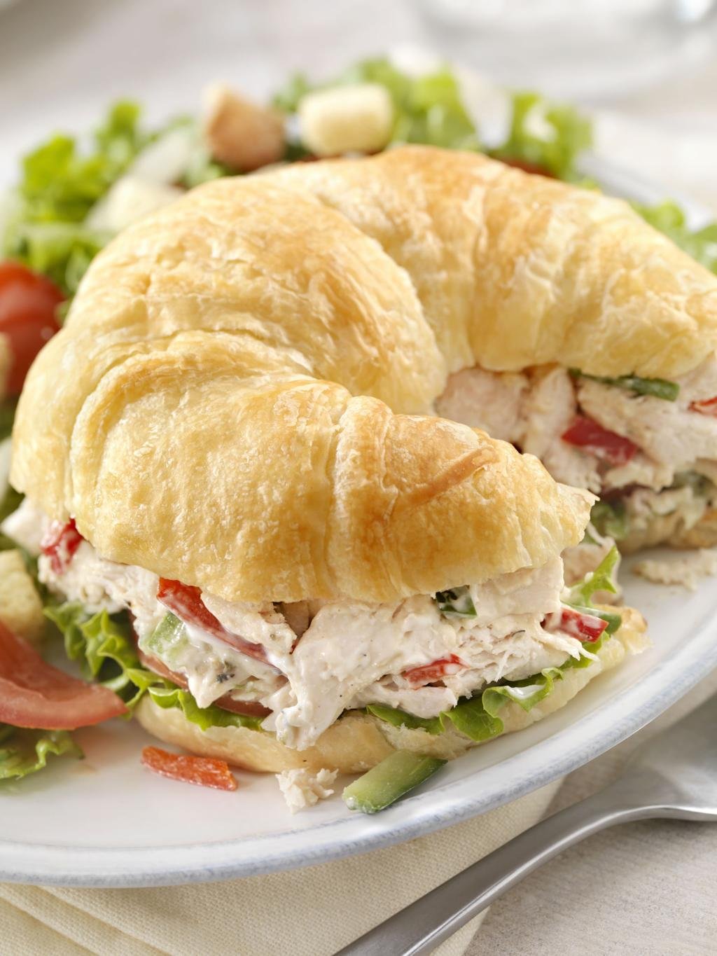 Croissant sandwich filled with chicken salad, lettuce, and tomato on a plate with greens in the background