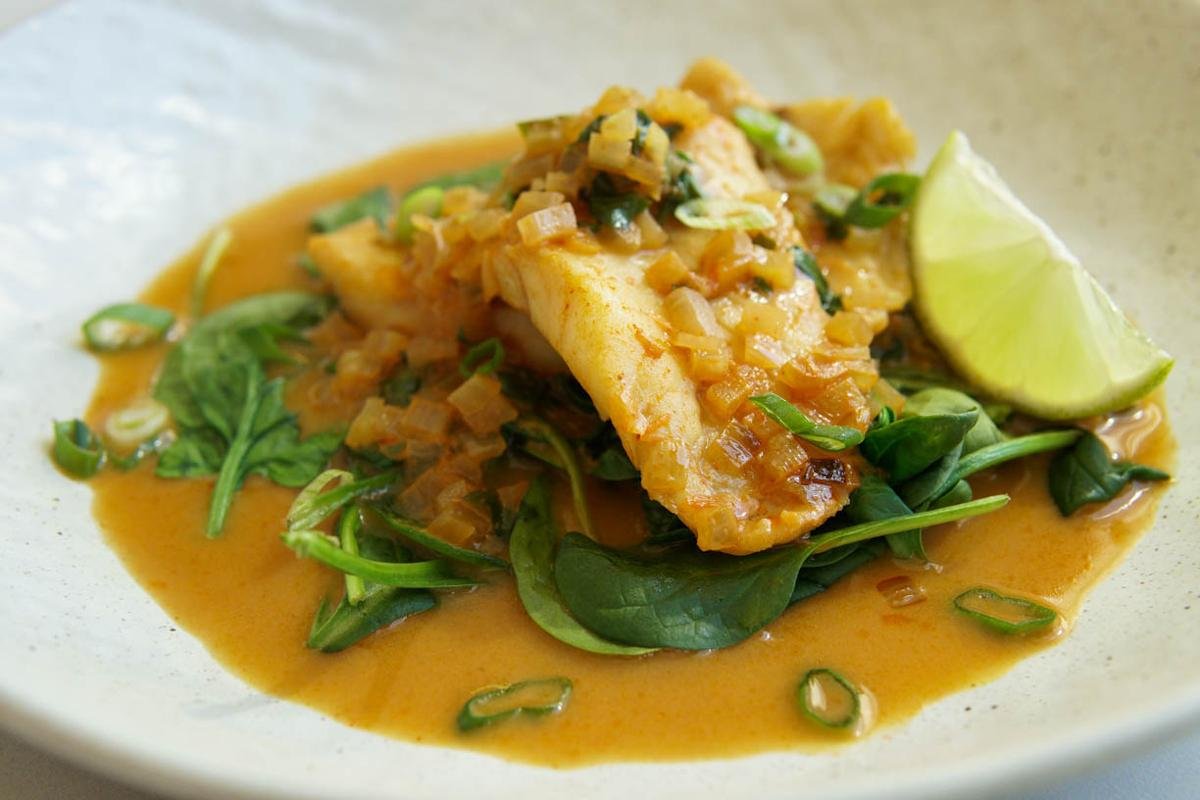 Piece of fish in orange sauce on a bed of greens with a lime wedge on the side, garnished with chopped onions and herbs.