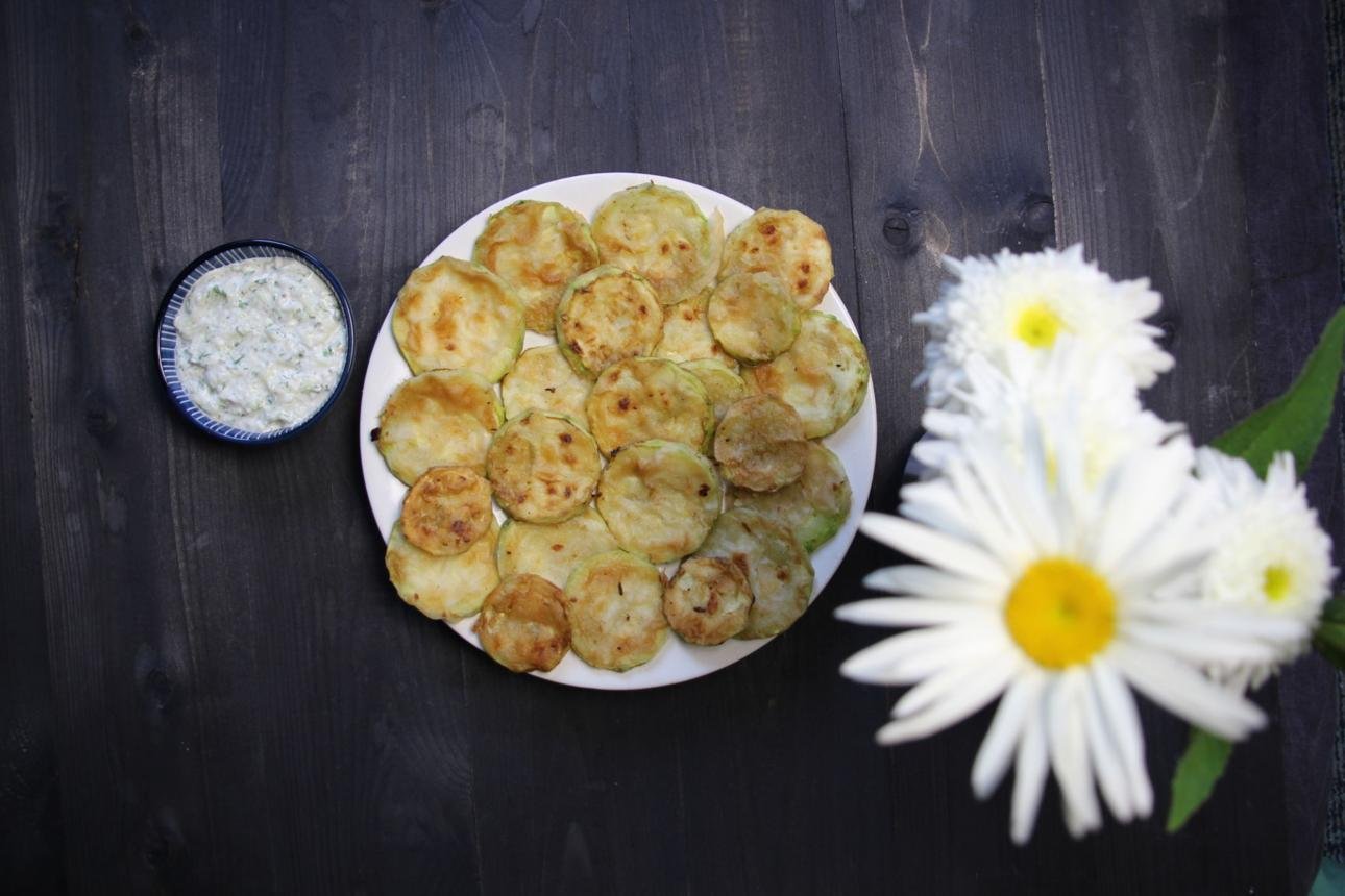 Plate of golden-brown zucchini fritters with a herb dip in a small blue bowl on a dark wooden table, with white daisies on the right.