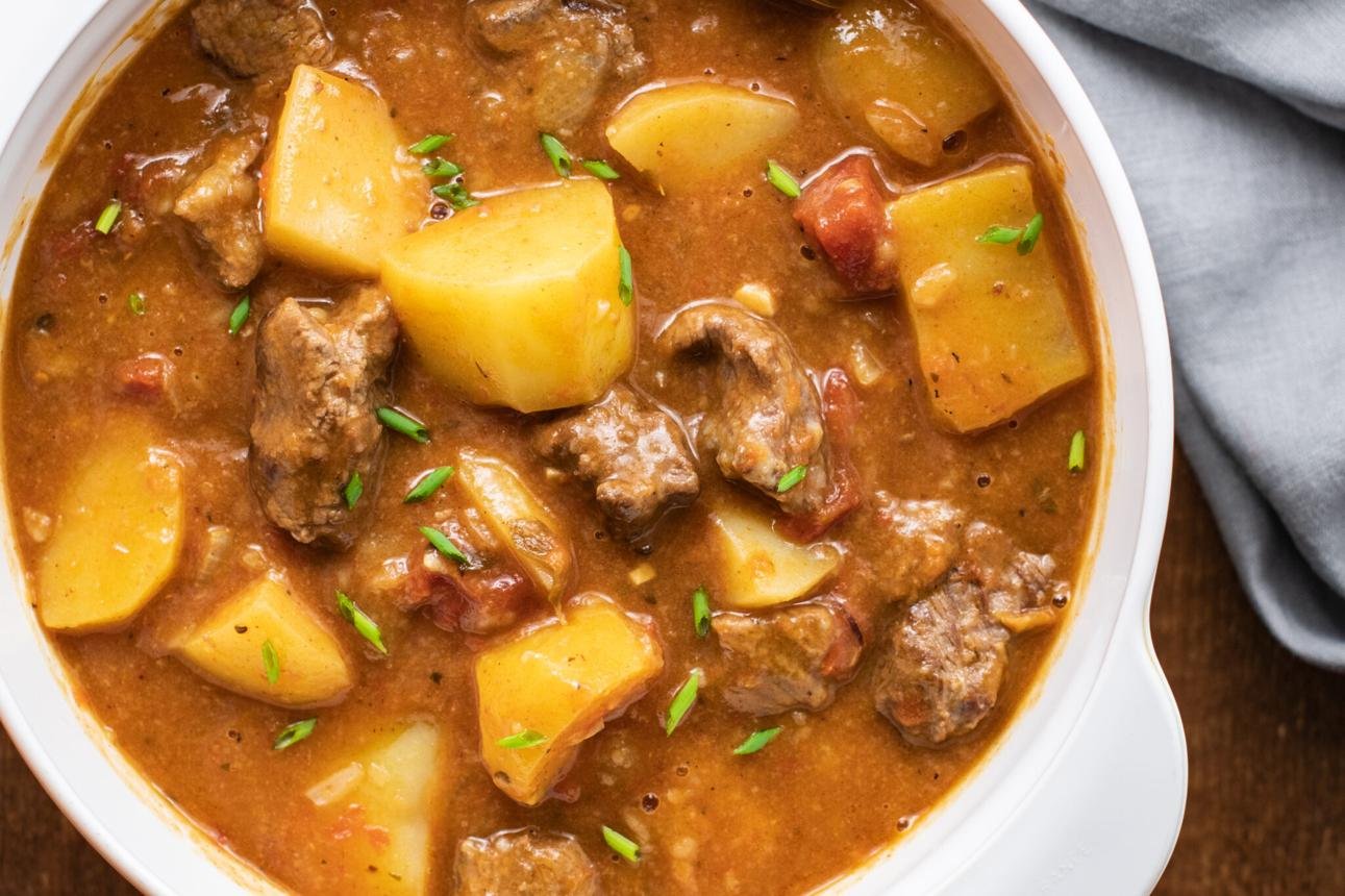 Beef and potato stew in a white bowl with chunky potatoes and tender meat in a tomatoy broth, garnished with green herbs.
