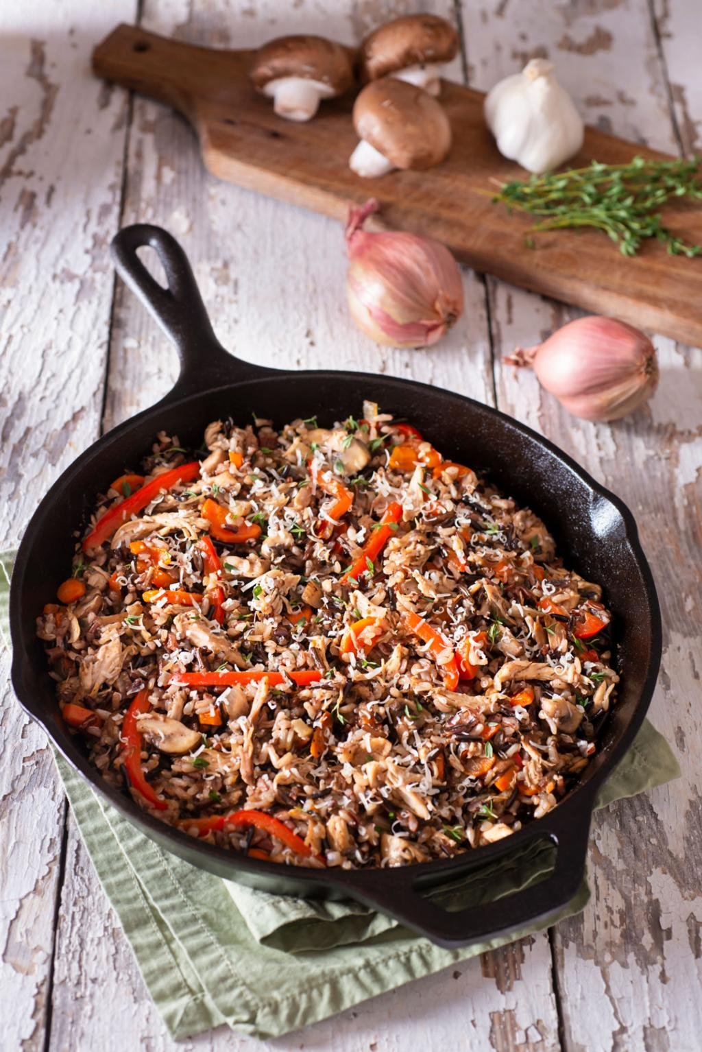 Skillet of mushroom, red pepper, and herb fried rice on a rustic wooden table, with garlic and thyme in the background.