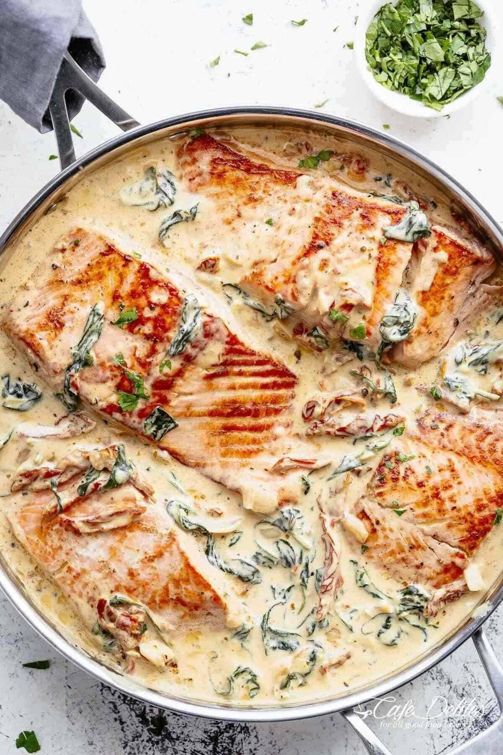 Salmon fillets in a creamy dill sauce with spinach in a skillet, topped with herbs and a bowl of chopped greens nearby.