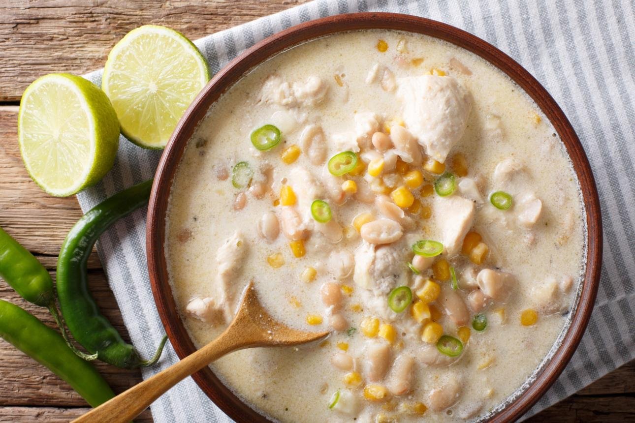 Creamy chicken soup with corn, beans, and chicken chunks in a brown bowl, garnished with sliced green onions on a striped cloth.