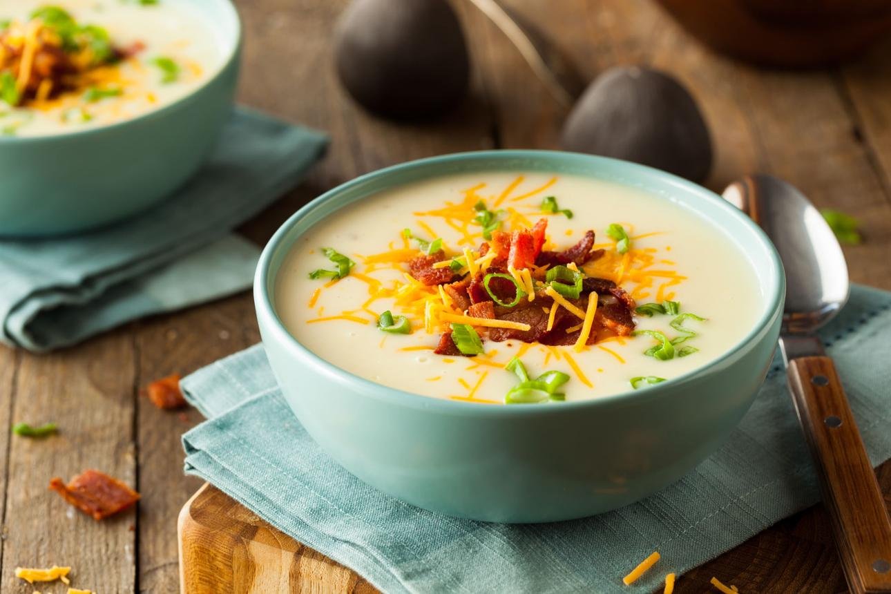 Creamy potato soup in a turquoise bowl topped with shredded cheddar, crispy bacon, and chopped green onions on a wooden table.