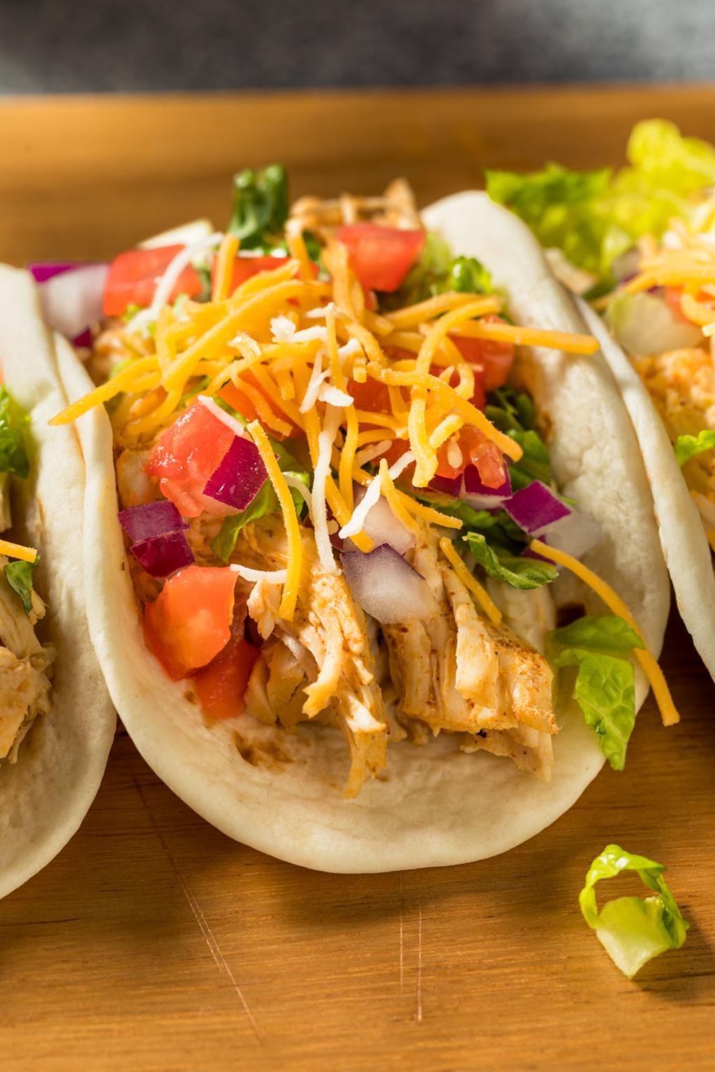 Soft flour tortillas filled with shredded chicken, lettuce, tomatoes, red onion, and cheddar cheese on a wooden surface.