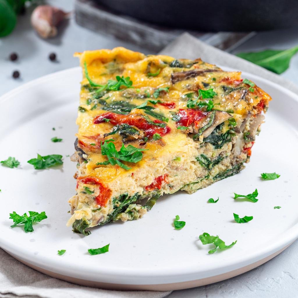 A colorful vegetable frittata slice with tomatoes, greens, and herbs on a white plate, garnished with parsley.