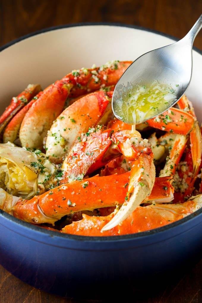 Snow crab legs coated in garlic herb butter being poured over in a blue pot with a spoon above. (informative)
