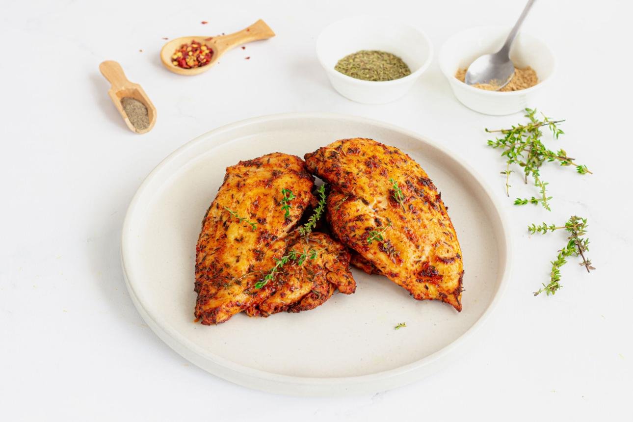 Herb-seasoned grilled chicken breasts on a white plate with thyme sprigs and small bowls of spices in the background.