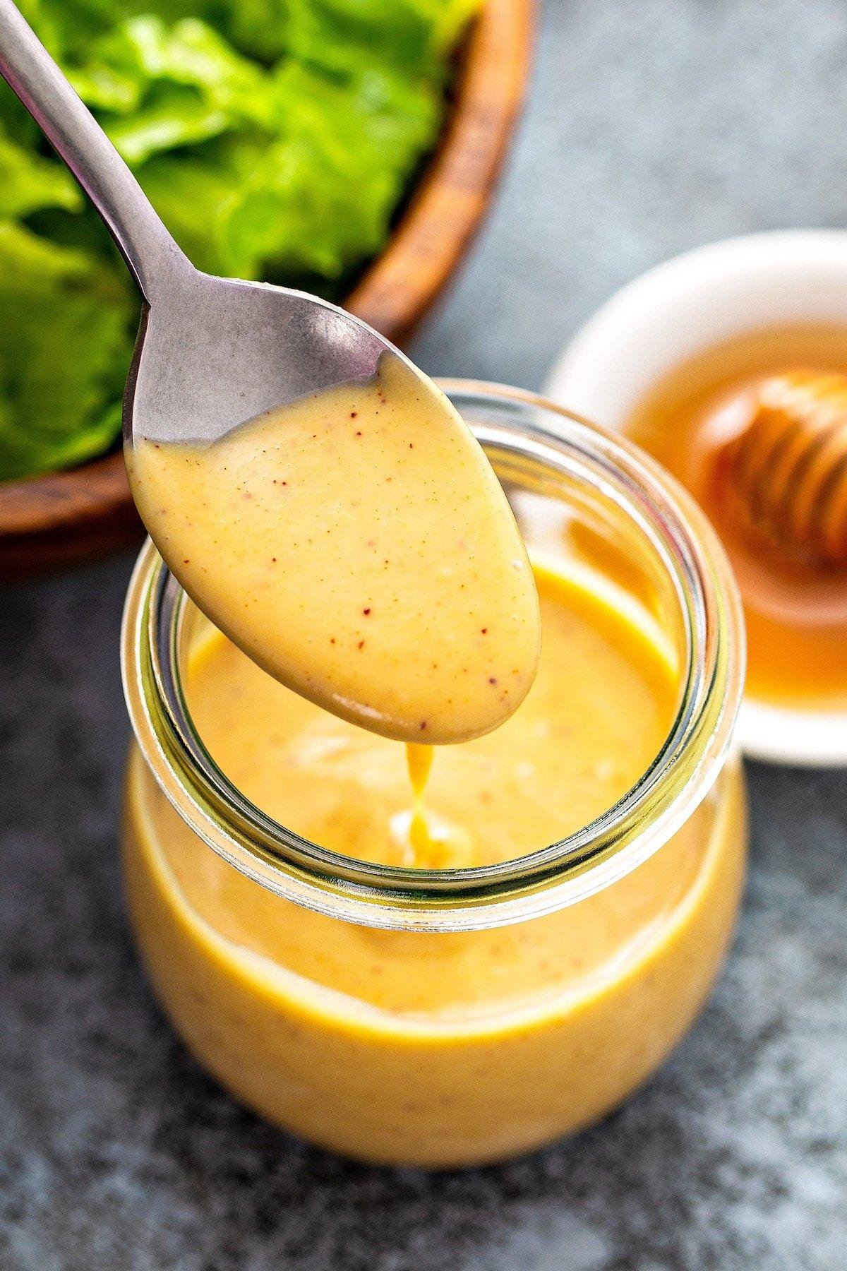 Creamy honey mustard sauce with mustard seeds on a spoon above a glass jar, honey and greens in the background