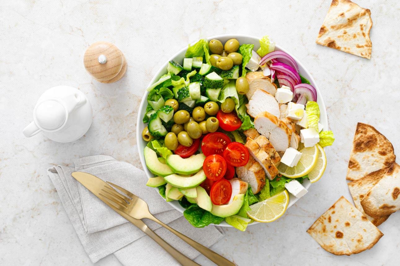 Large bowl of mixed salad with grilled chicken, avocado, olives, cucumbers, tomatoes, feta, lettuce, and lemon wedges; pita breads on the side.