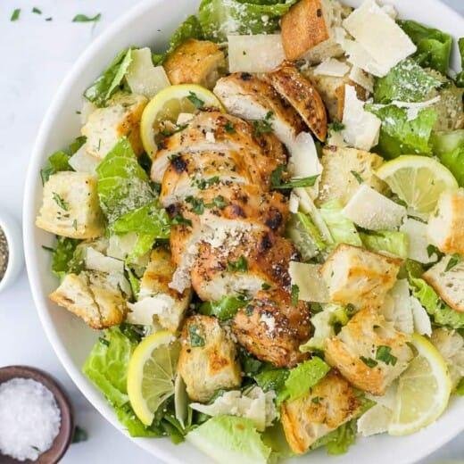 Grilled chicken slices on a Caesar-style salad with lettuce, croutons, shaved parmesan, and lemon wedges.