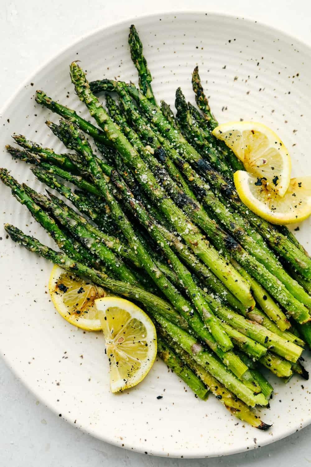 Roasted green asparagus spears arranged on a white plate with lemon wedges and a sprinkle of salt and pepper.