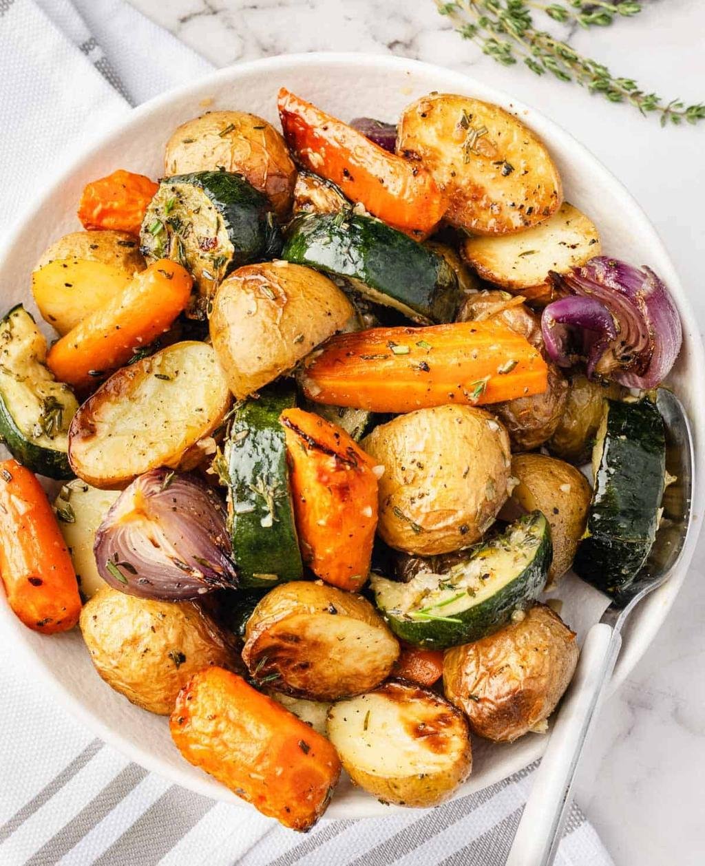 Roasted potatoes, carrots, zucchini, and red onions seasoned with herbs in a white bowl.