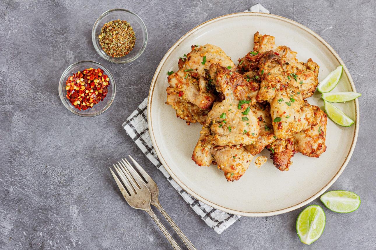 Plate of crispy fried chicken wings garnished with chopped herbs, with lime wedges on the side and small bowls of crushed red pepper and spices nearby on a gray surface.