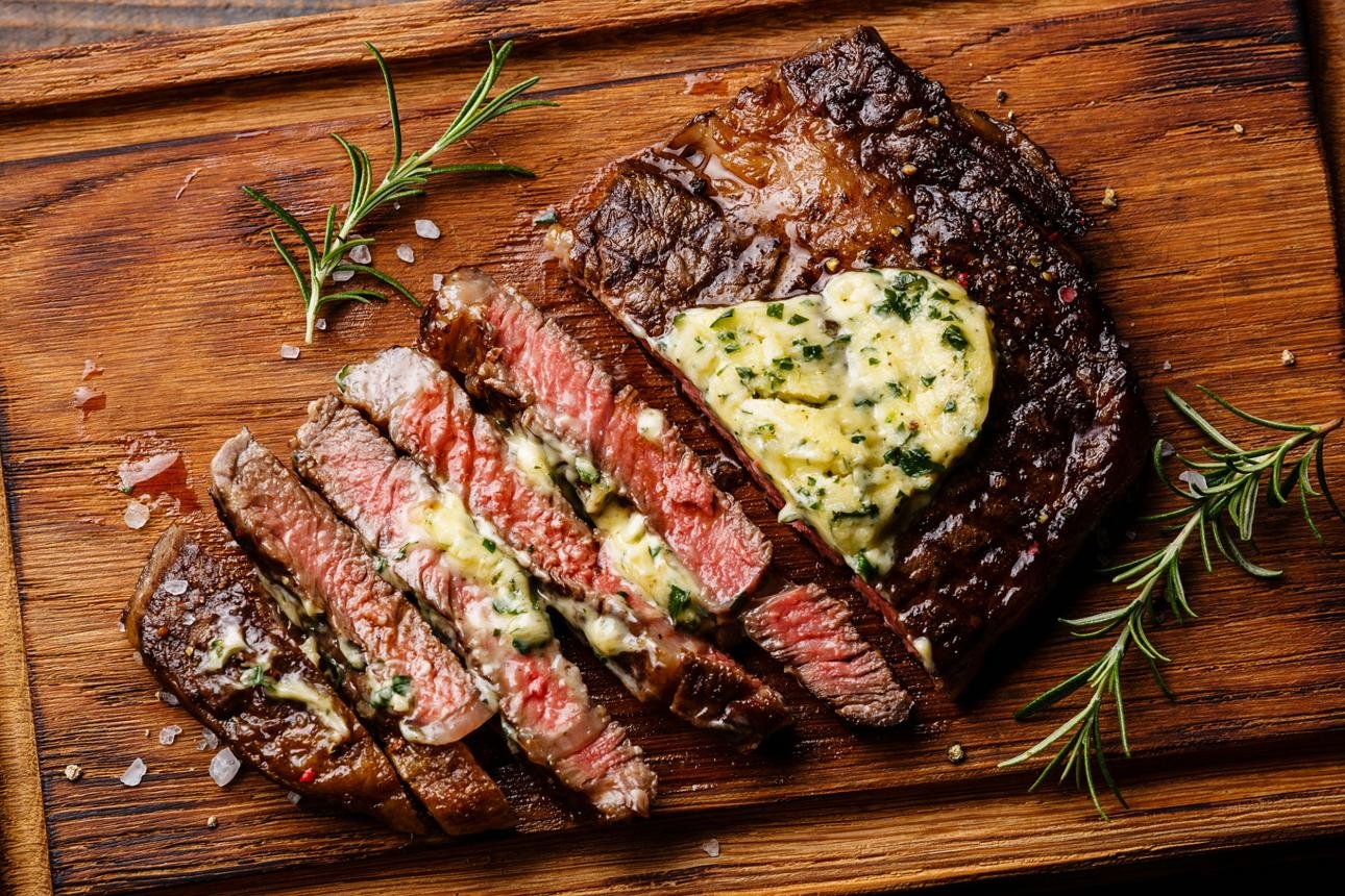 Sliced medium-rare steak on a wooden board with garlic-herb butter and rosemary sprigs