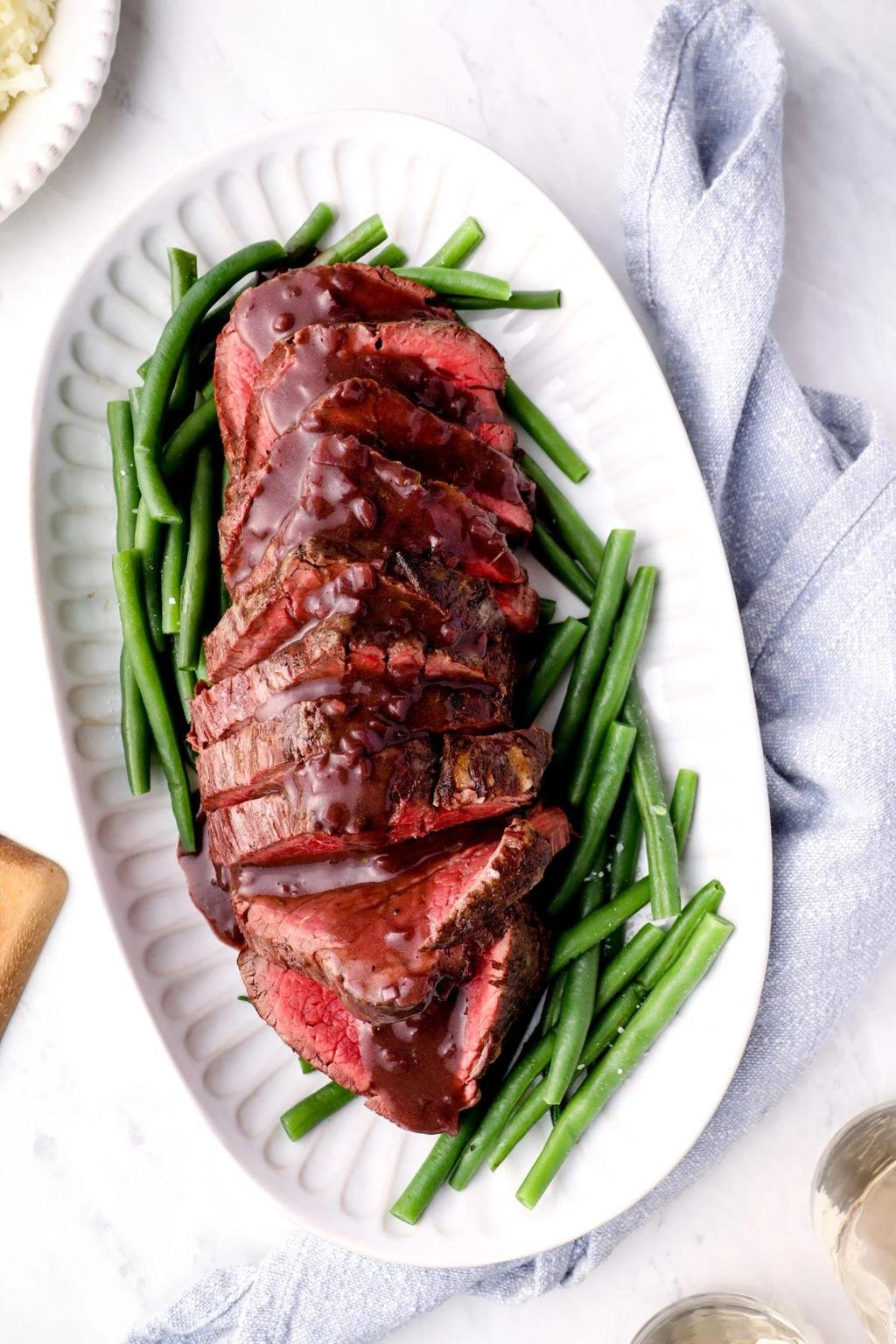 Sliced roast beef with a dark glaze served over green beans on a white platter.