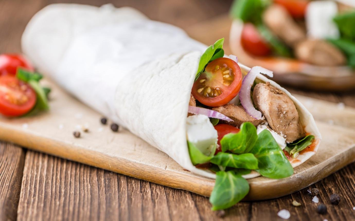 Tortilla wrap filled with grilled chicken, cherry tomato, red onion, greens, on a wooden cutting board