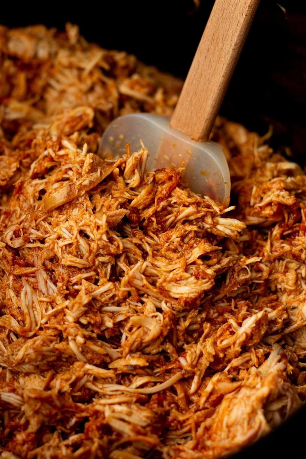 Shredded pulled pork being stirred in a pan with a wooden spatula, glossy with barbecue sauce.
