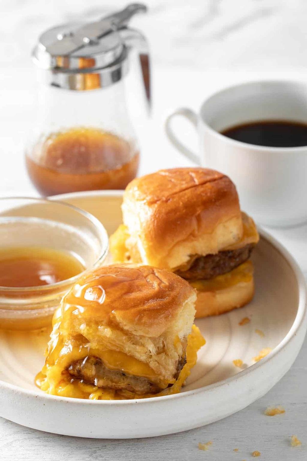 Two cheese-stuffed sliders on a white plate with maple syrup, a coffee cup, and a glass syrup pitcher in the background.