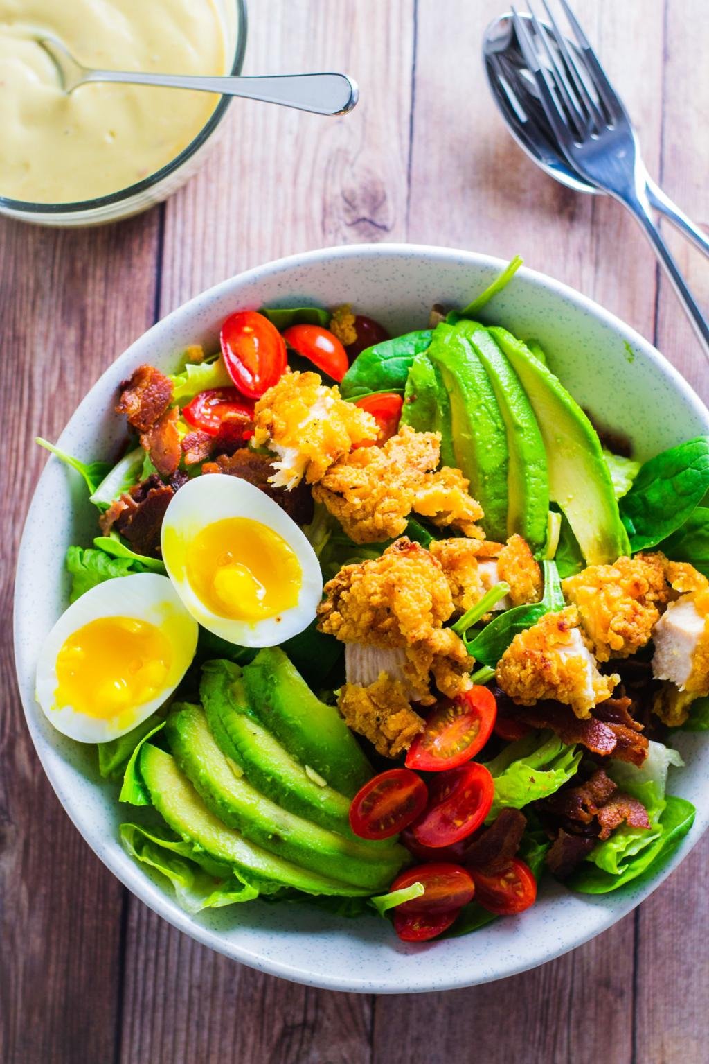 Bowl of salad with avocado slices, soft-boiled eggs, cherry tomatoes, greens, and crispy fried chicken topping