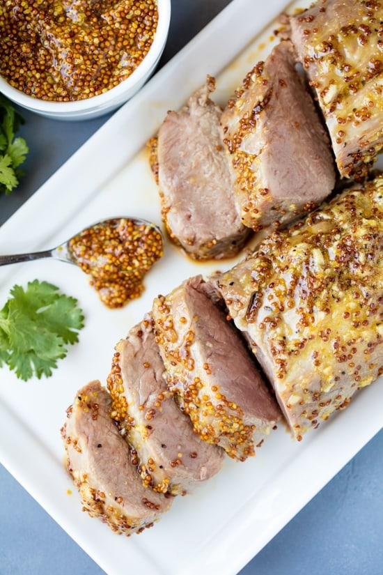 Sliced roasted pork tenderloin with a mustard seed crust on a white serving platter, with a spoonful of mustard nearby.