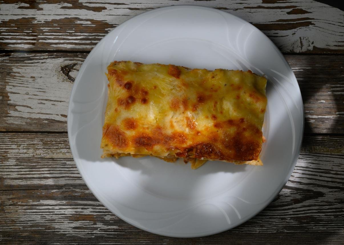 A cheesy lasagna slice on a white plate, placed on a rustic wooden table from a top-down view.