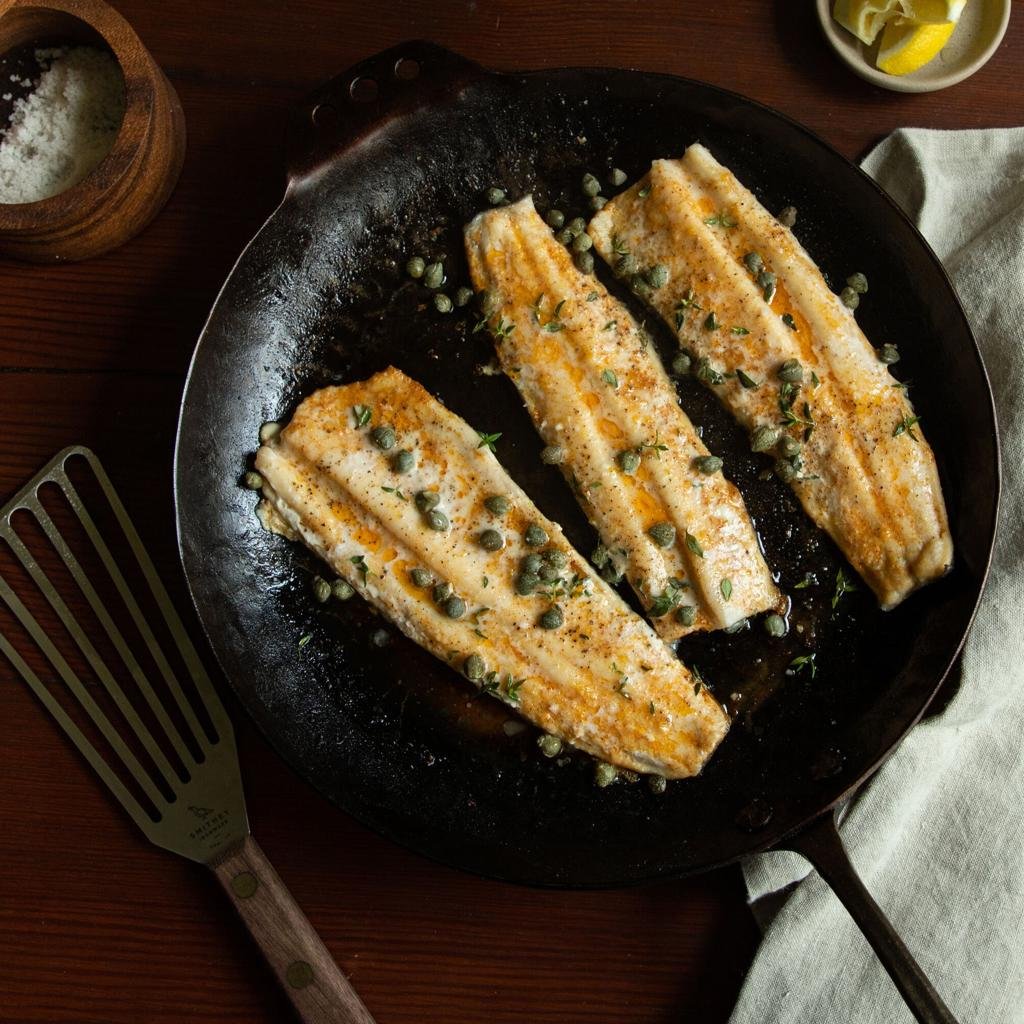Three pan-seared white fish fillets sprinkled with capers and herbs in a black skillet on a wooden table
