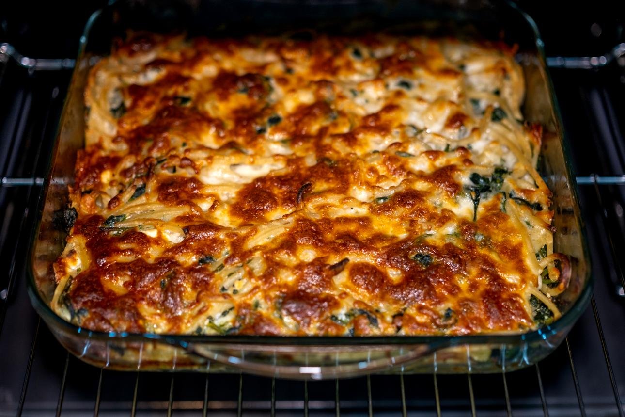Cheesy spinach and noodle casserole baking in a glass dish inside an oven, with a golden-brown crust on top.