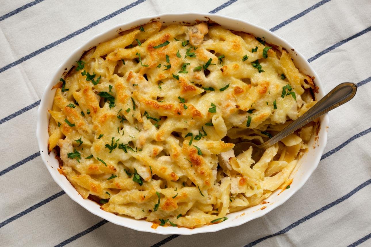 Baked macaroni and cheese in a white oval dish, topped with a golden crust and chopped parsley, with a serving spoon inside.