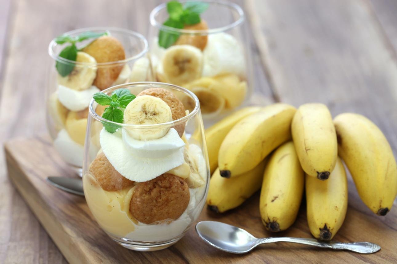 Parfait glasses with banana, cream, and cookie crumbles, garnished with mint on a wooden board.