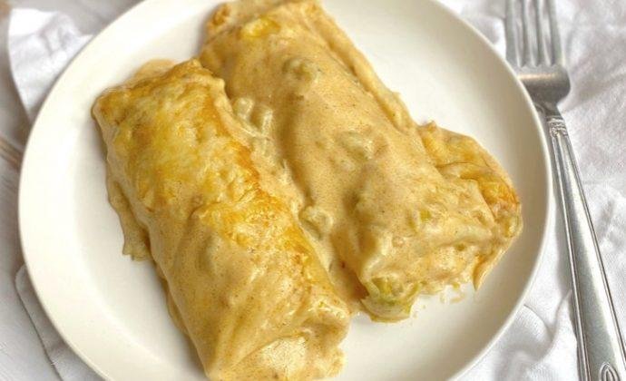Two rolled tortillas on a white plate smothered in a light cream sauce, served with a fork nearby.