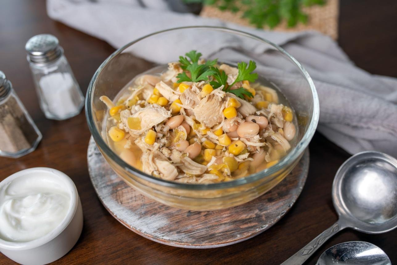 Bowl of shredded chicken, corn and white beans in broth garnished with parsley on a wooden board. Nearby yogurt, salt and pepper shakers, and metal spoon visible.