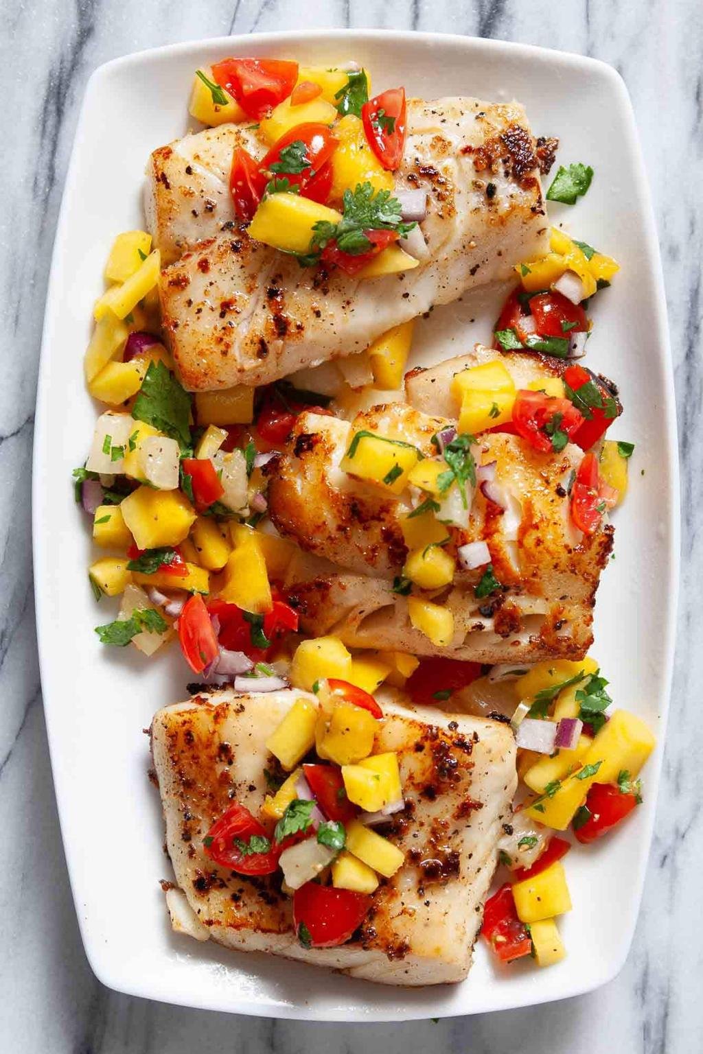 Pan-seared white fish fillets with mango salsa (diced mango, red pepper, onion, cilantro) on a white platter beneath a marble backdrop.
